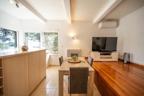A modern kitchen with a wooden island, a dining table with chairs, a built-in TV, and a large window offering a view of greenery.