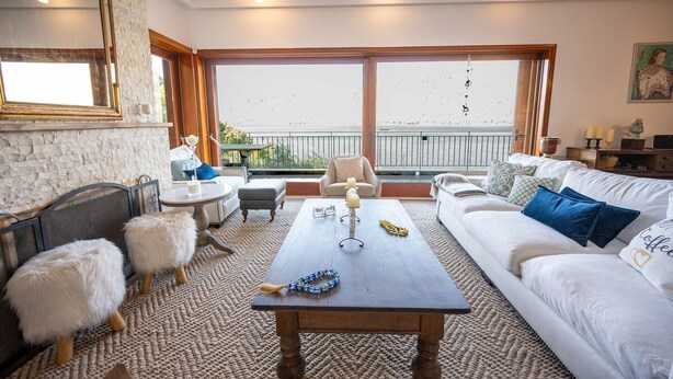 Ein gemütliches Wohnzimmer mit einer Steinwand, einem Holztisch, einem weißen Sofa, weichen weißen Poufs und Meerblick durch große Fenster.