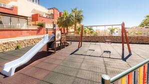 Un terrain de jeux avec un toboggan et des balançoires, situé devant un décor composé d’immeubles et de palmiers.