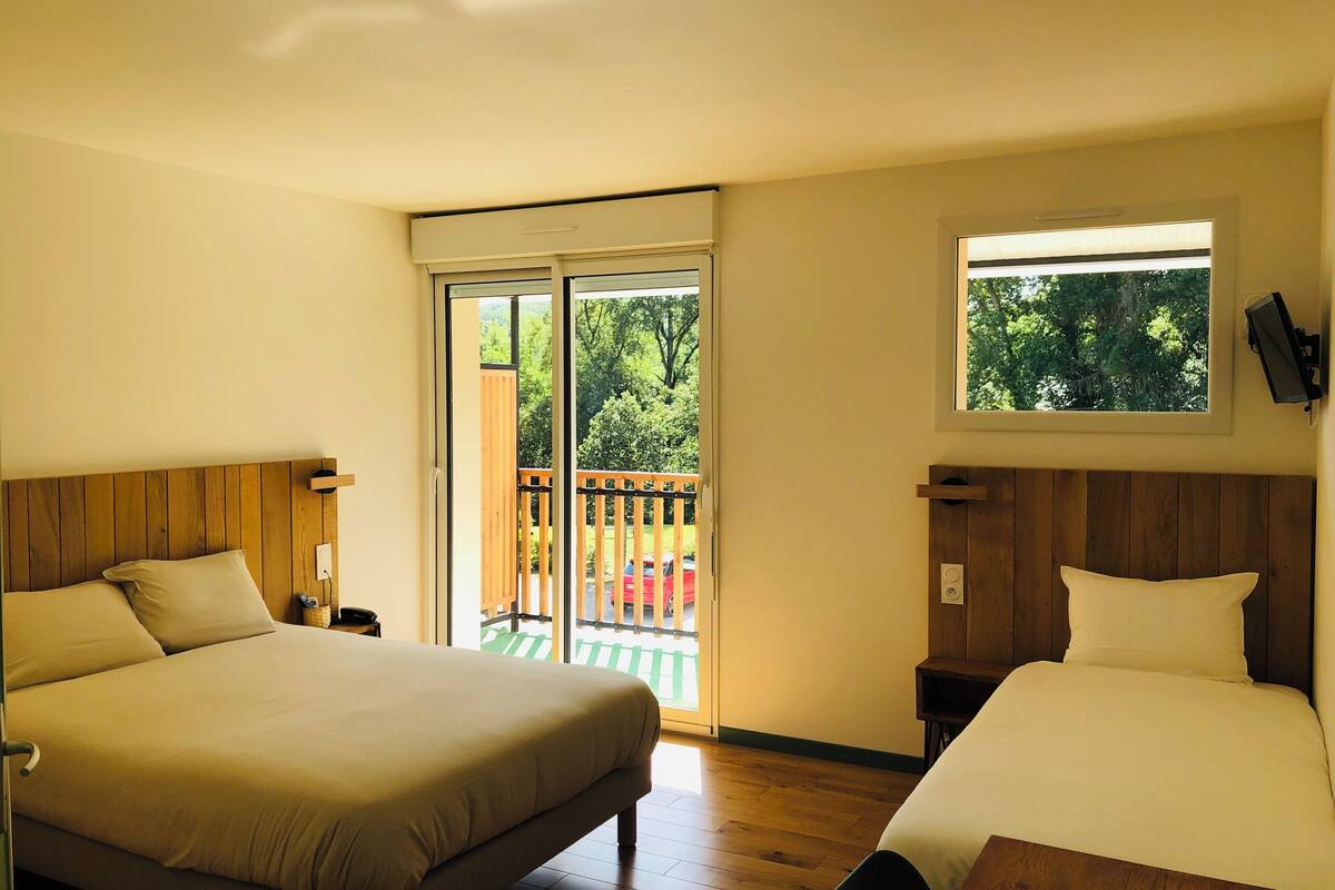 Habitación de hotel con dos camas, suelo de madera, balcón con vista a árboles y ventana con vista al exterior.