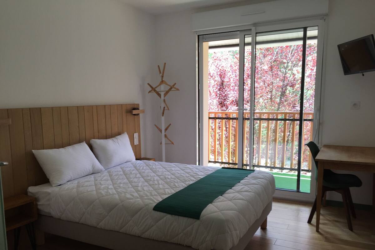 Un dormitorio con cama, cabecera de madera, manta verde, mesita de noche de madera, escritorio, silla y ventana con vista a árboles.