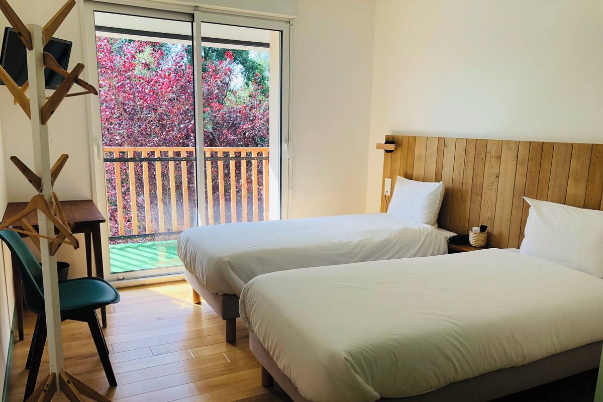 Habitación de hotel con dos camas, cabecero de madera, escritorio con silla y balcón con vista a árboles y una cerca.