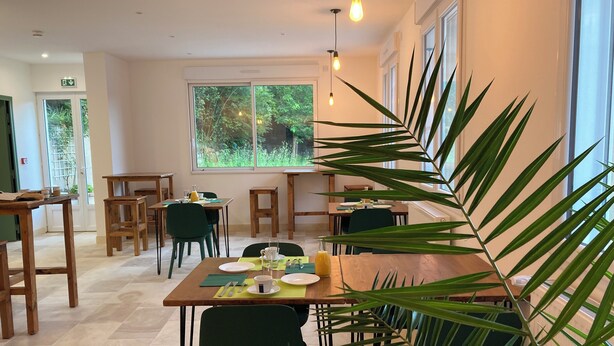 Comedor con mesas y sillas de madera, un ventanal con vistas a follaje e iluminación colgante.