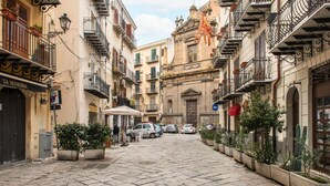 Un carrer empedrat i estret flanquejat per edificis de diverses plantes, amb balcons amb plantes, un cafè i cotxes aparcats.