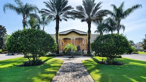 A building with a paved pathway leading to a covered entrance, surrounded by well-maintained grass and palm trees. - Modern 5BR Retreat w/ Pool and Clubhouse Access (Kissimmee)
