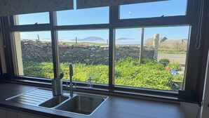 A kitchen with a large window offering a view of a stone wall, greenery, and distant mountains. - Withi-gill - Bay of Ireland Views 50m From Beach (Stromness)