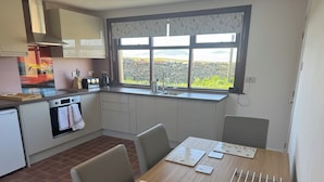 A modern kitchen with a dining area, featuring a large window with a view of greenery and mountains, a built-in oven, and a dining table with place settings. - Withi-gill - Bay of Ireland Views 50m From Beach (Stromness)