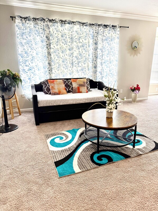Un salon comprenant un canapé, une table basse ronde, un tapis à motifs, un ventilateur et un vase avec des fleurs.