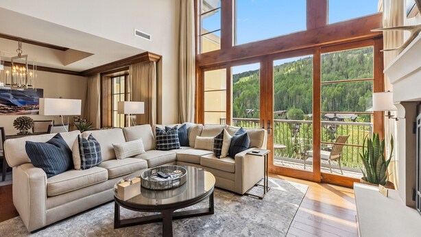 A spacious living room with a large sectional sofa, a coffee table, and a view of a forested area through large windows. - Rare Two Story Condo in Ritz-Carlton by InvitedHome | Pool, Hot Tub, Lounge, Gym (Vail)