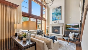 A spacious living room with a fireplace, large windows, and a modern chandelier. - Rare Two Story Condo in Ritz-Carlton by InvitedHome | Pool, Hot Tub, Lounge, Gym (Vail)