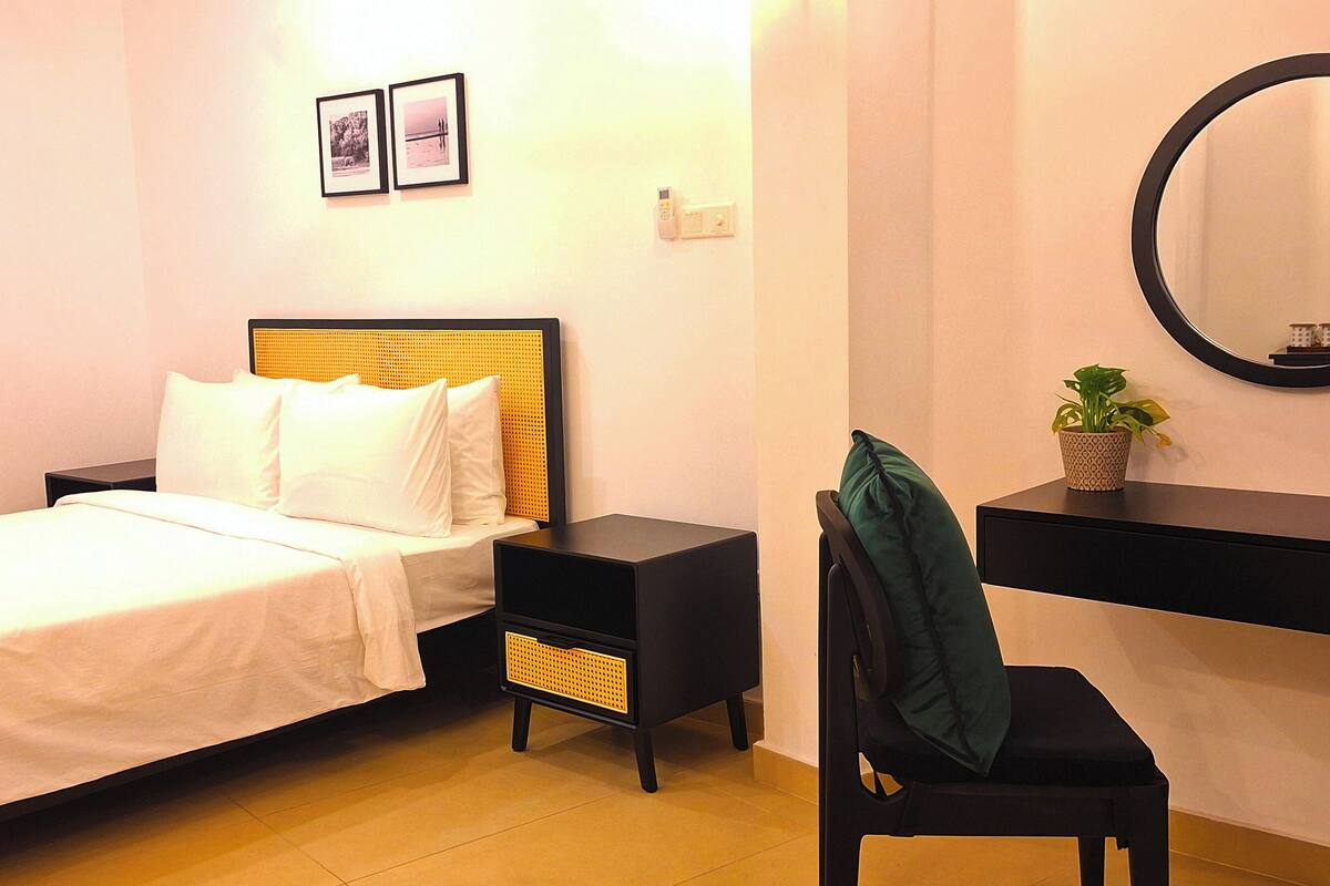 Un dormitorio con cama, mesita de noche, silla, escritorio, espejo y estante de pared con una planta.