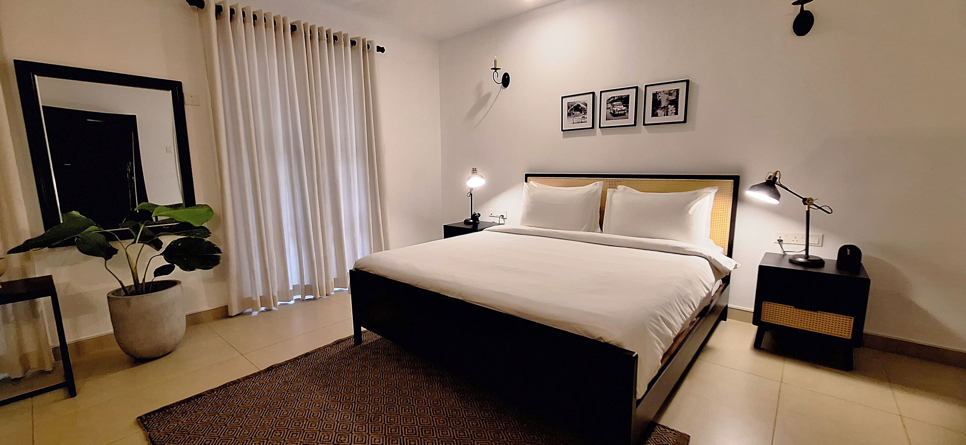 A modern bedroom with a large bed, a potted plant, a nightstand with a lamp, and a wall-mounted shelf.