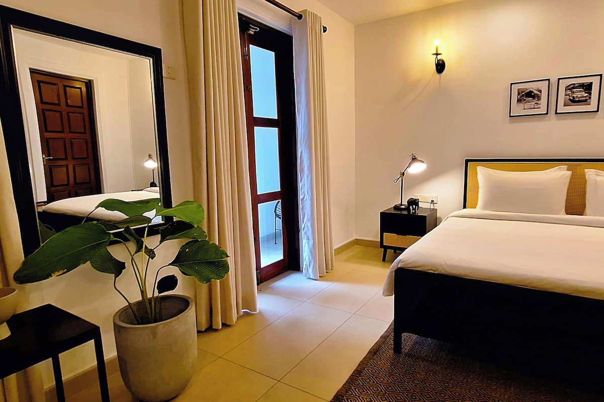 Una habitación de hotel con cama, mesita de noche, lámpara, planta en maceta y una puerta que da a otra habitación.