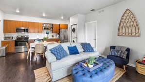 A modern kitchen with stainless steel appliances, a dining area with a striped sofa and blue cushions, and a living area with a blue ottoman and a decorative wall piece. - Cozy Nautical Themed 2-bedroom condo with AC in charming Traverse City. (Traverse City)