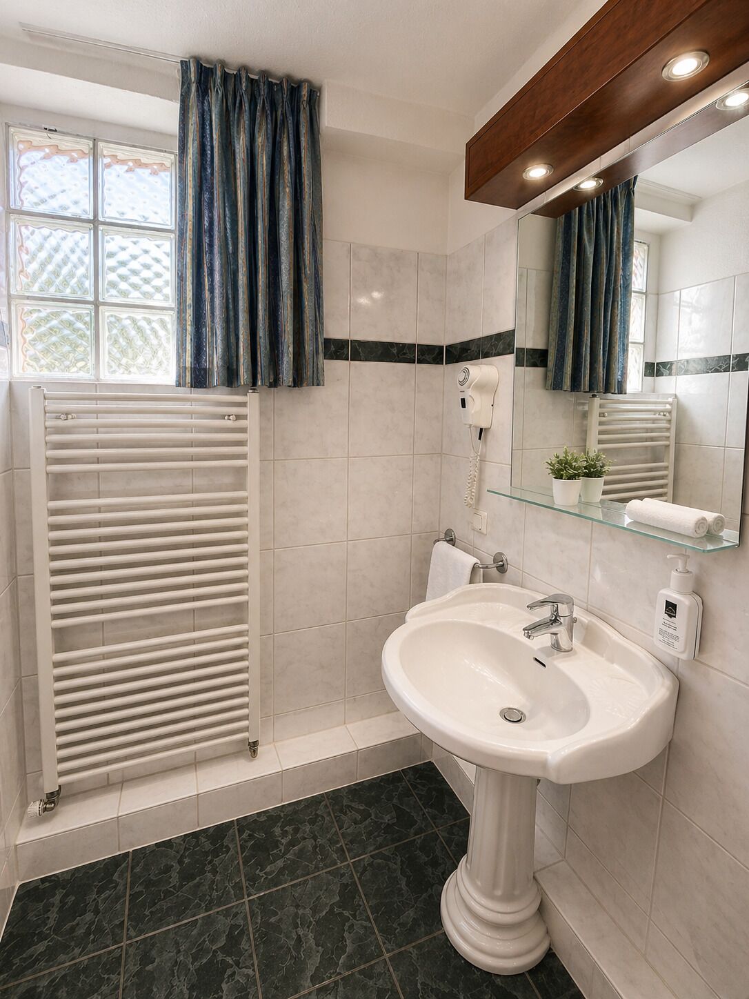 A bathroom with a white sink, a glass shelf with toiletries, a towel rack, a mirror, and a window with curtains.
