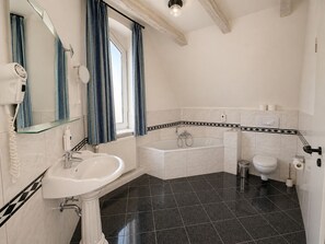 A bathroom with a sink, mirror, window with curtains, bathtub, toilet, and a shower area.