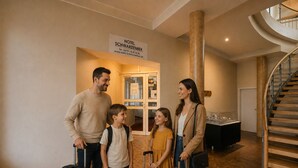 Une famille, avec des valises, se trouve dans le hall d’un hôtel, près d’un escalier et d’un panneau indiquant l’hôtel Schwarzenbek.