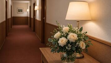 Un pasillo con un banco de madera, un jarrón de vidrio transparente con flores blancas y una lámpara con pantalla color beige.