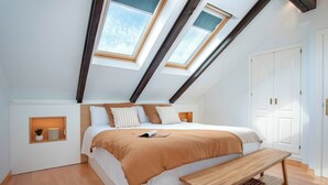 A bedroom with a sloped ceiling, skylights, a bed with a brown blanket, a bedside table, and a wooden bench.