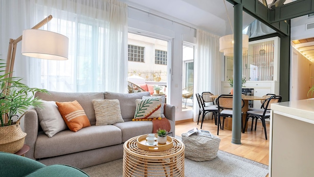 A modern living room with a grey sofa, a round wicker coffee table, and a dining area with wooden chairs and a white table.
