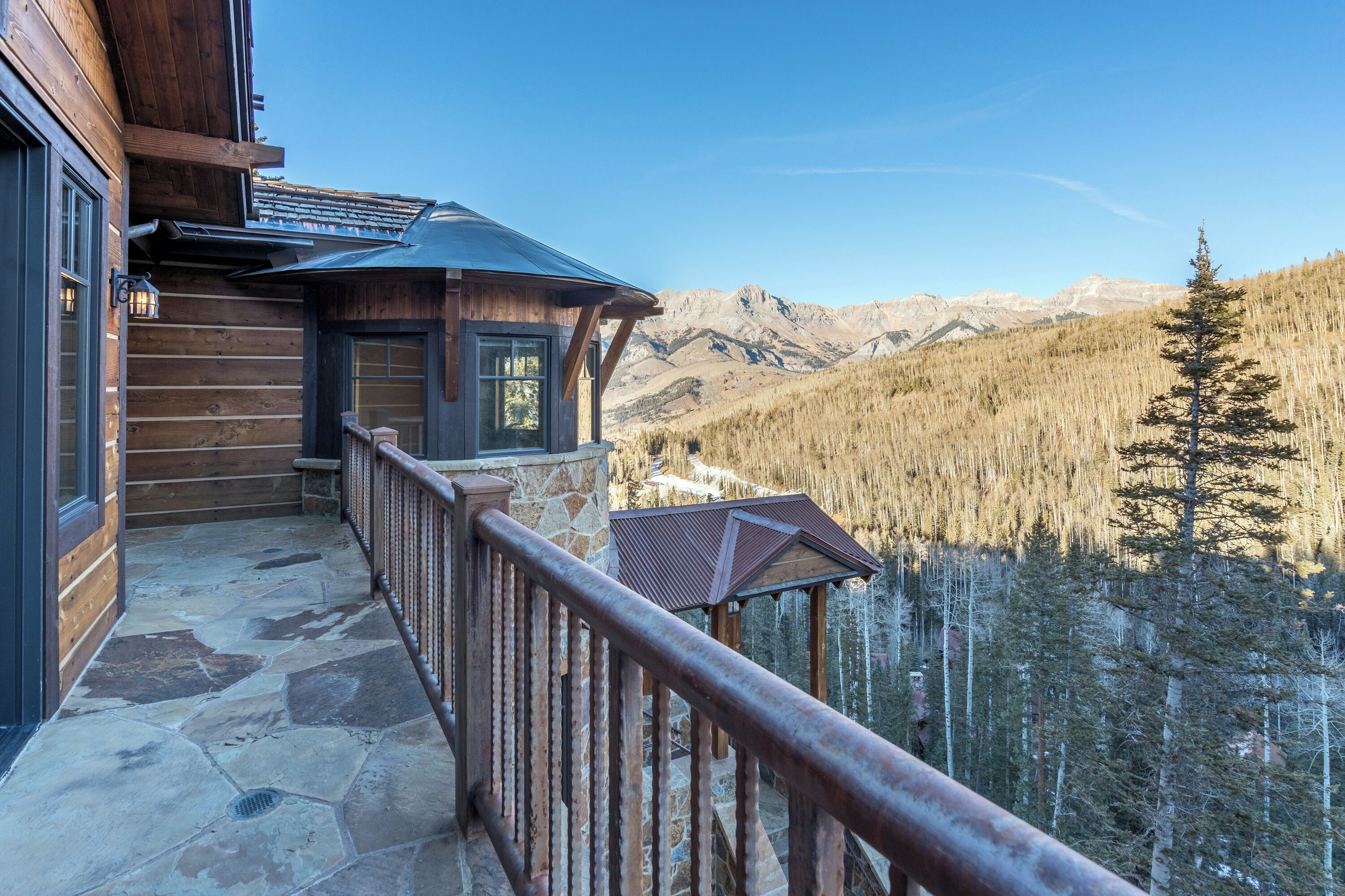 A wooden cabin with a stone-paved balcony overlooking a forested valley and mountains.