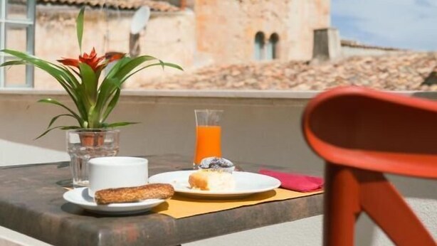 A table with breakfast items including a cup, a glass of orange juice, a pastry, and a plant. In the background, there is a stone building with a tower. - U Zuccareddu (Alcamo)