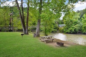 Un área de picnic con banca y mesa, junto a un río, rodeada de árboles y un puente de madera.