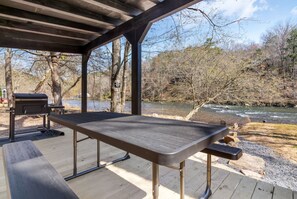 Zona de picnic al aire libre con mesa y parrilla, con vistas a un río y a zona boscosa.