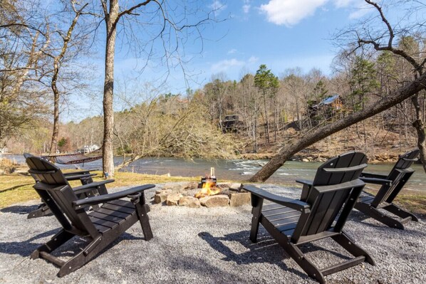 Un camping con tres sillas Adirondack, una fogata y vistas a un río y bosque.