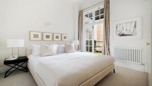 A bedroom with a large bed, a bedside table with a lamp, and a window with curtains. - Elegant 3-Bedroom Luxury Retreat in Prestigious Cadogan Square (London)