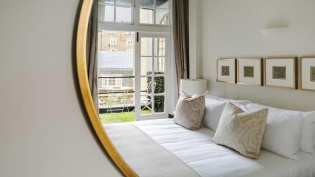 A hotel room with a white sofa, a large window with a view of buildings, and framed artwork on the wall. - Elegant 3-Bedroom Luxury Retreat in Prestigious Cadogan Square (London)