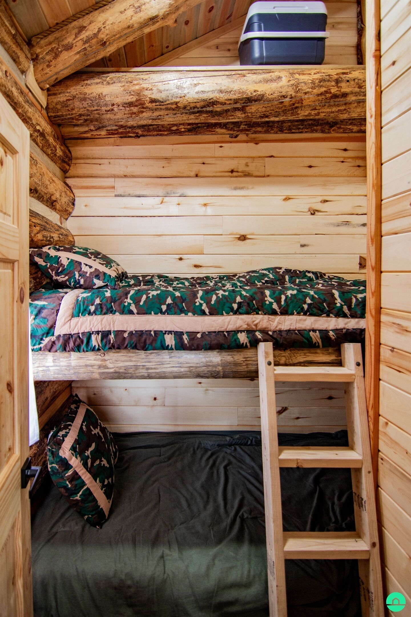 Una litera con ropa de cama estilo camuflaje, un pequeño box de almacenamiento en la parte superior y una escalera de madera.
