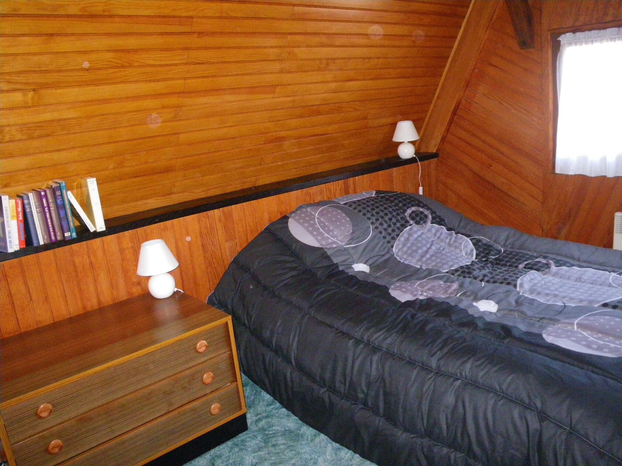 A bedroom with a wooden bunk bed, a bedside table with a lamp, and a shelf with books.