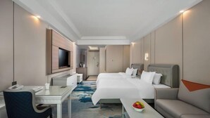 A hotel room with a bed, a desk, a chair, a TV, and a small table with fruit. - Jun Yi Kai Di Hotel (Shenzhen International Convention and Exhibition Center) (Shenzhen)
