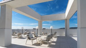 Een terras op het dak met witte stoelen en tafels, een helderblauwe lucht en een uitzicht op verre heuvels.