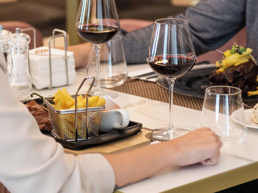 Una mesa de comedor con un bistec, papas fritas y un postre, junto con copas de vino y una cesta de fruta.