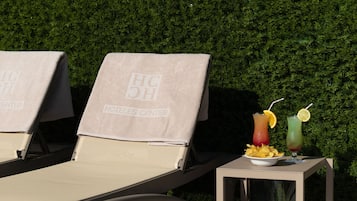 Two sun loungers with towels, a table with a drink and chips, and a hedge in the background.