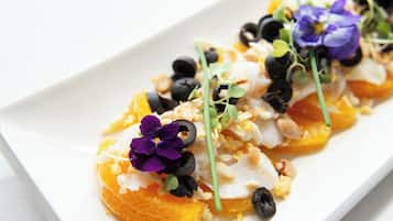 A white rectangular plate with a food arrangement featuring orange segments, purple flowers, black olives, and green sprouts.