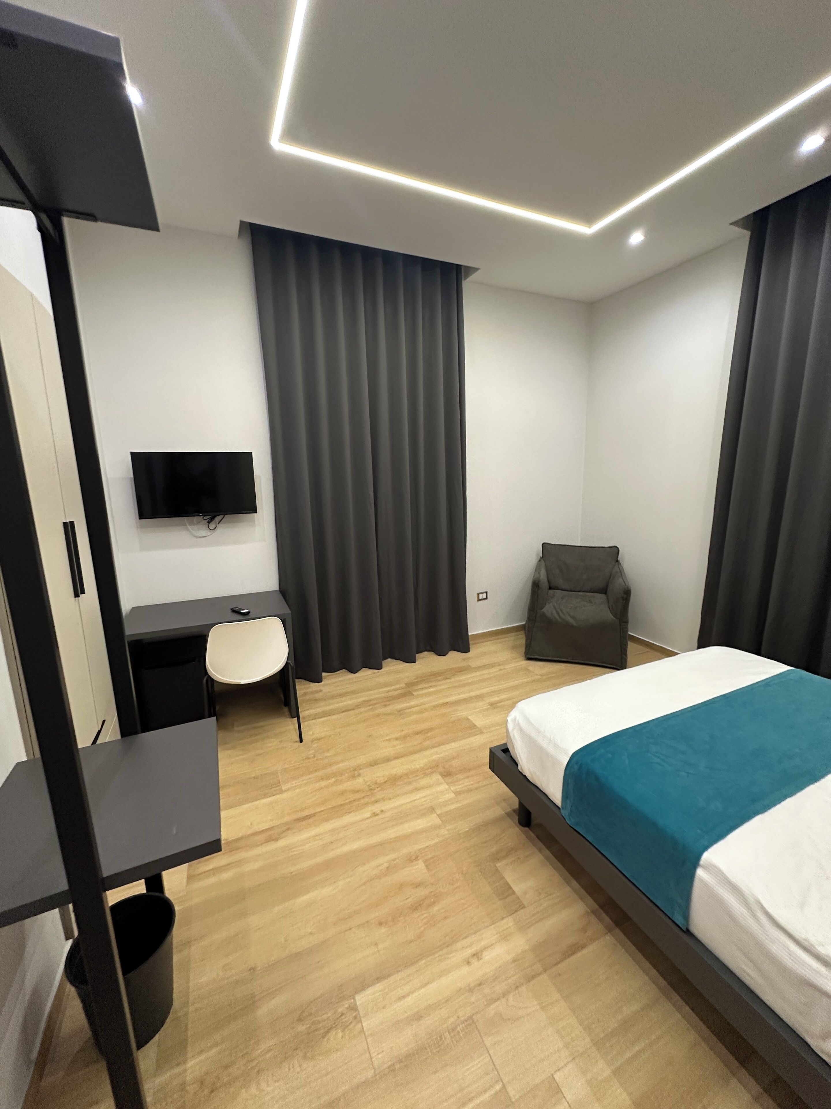 A modern hotel room with a bed, a desk, a chair, a TV, and a large window with curtains.