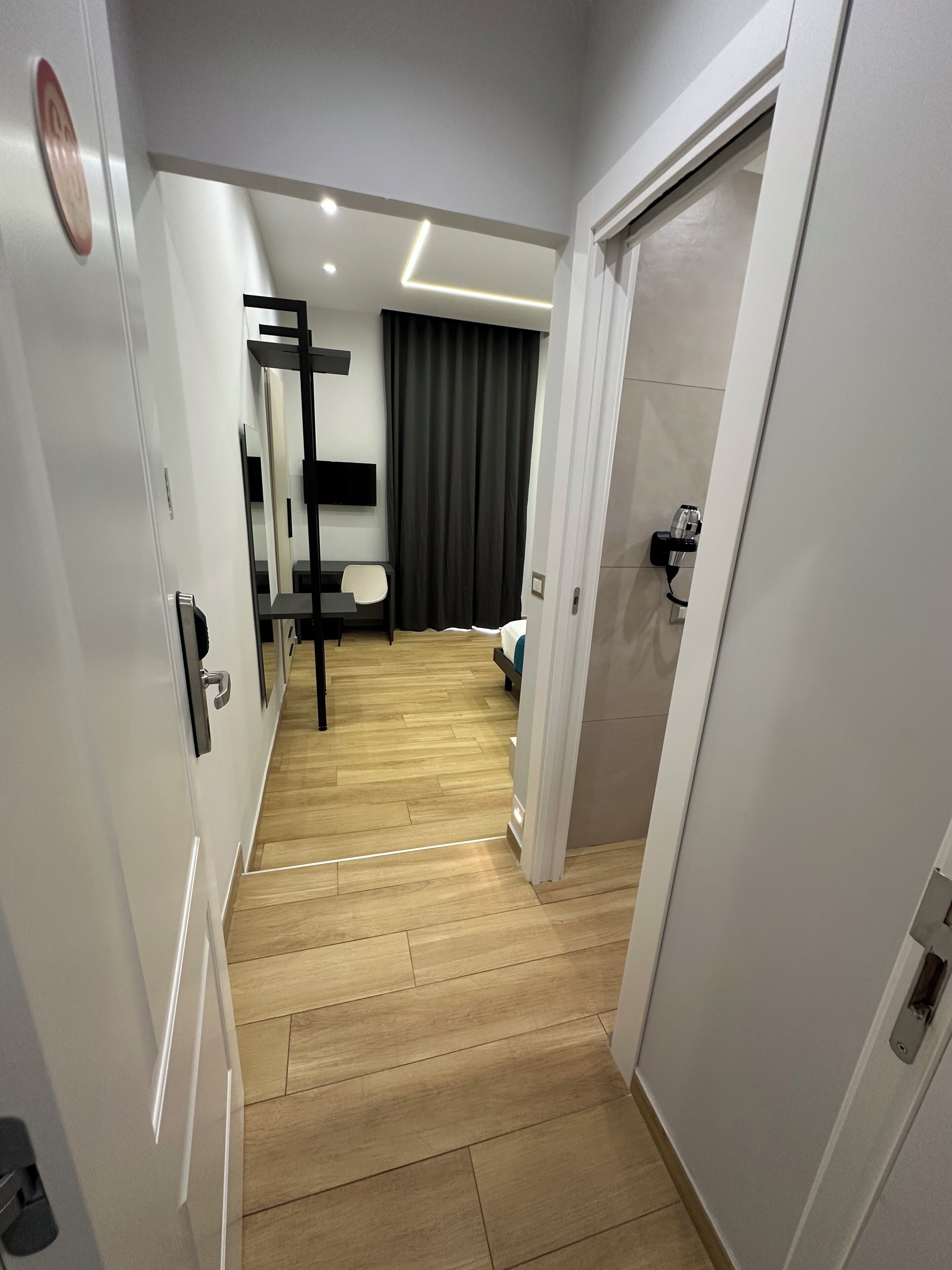 A modern hotel corridor with a wooden floor, a black chair, a bed, and a television on the wall.