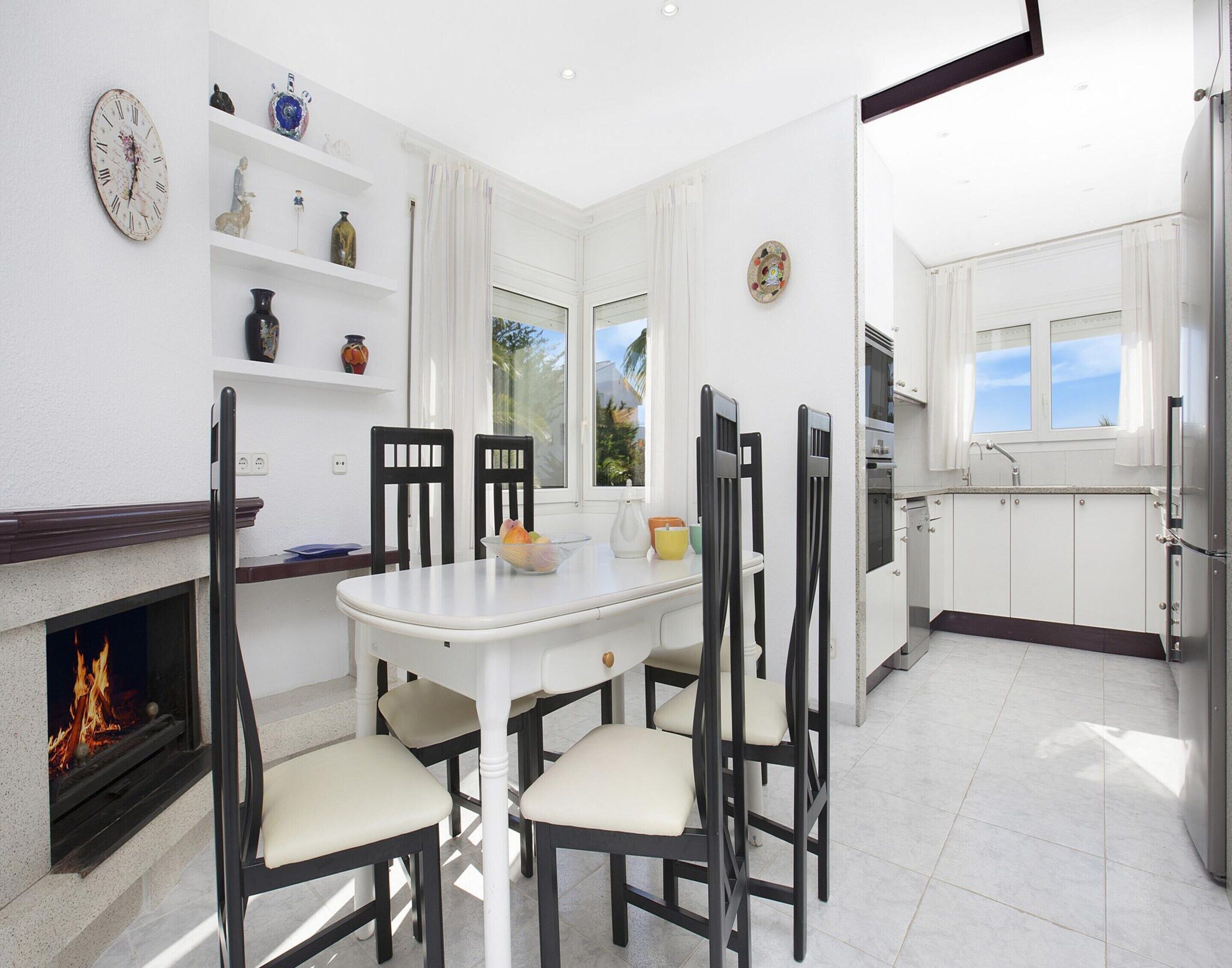 A modern kitchen with white cabinets, a dining table with chairs, a fireplace, and a view of the outdoors through the windows.