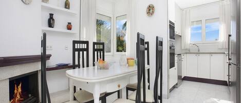 A modern kitchen with white cabinets, a dining table with chairs, a fireplace, and a view of the outdoors through the windows.