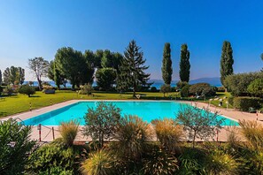 A rectangular swimming pool surrounded by well-maintained grass and trees, with a clear view of the sea in the background. - Sporting 2 58 by Wonderful Italy (Sirmione)