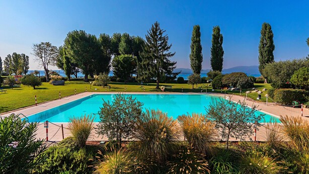 A rectangular swimming pool surrounded by well-maintained grass and trees, with a clear view of the sea in the background. - Sporting 1 64 by Wonderful Italy (Sirmione)