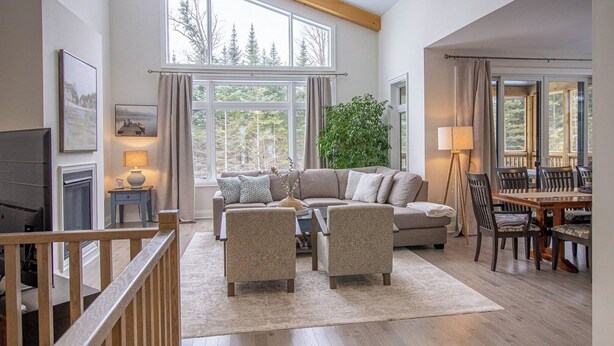 A spacious living room with a large window, a sofa set, a dining table with chairs, and a staircase.