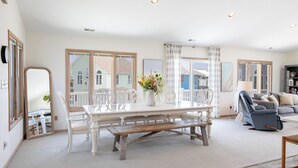A spacious living room with a large white dining table, white chairs, a grey sofa, and a grey recliner. There is a large window with curtains, a white floor lamp, and a bookshelf.