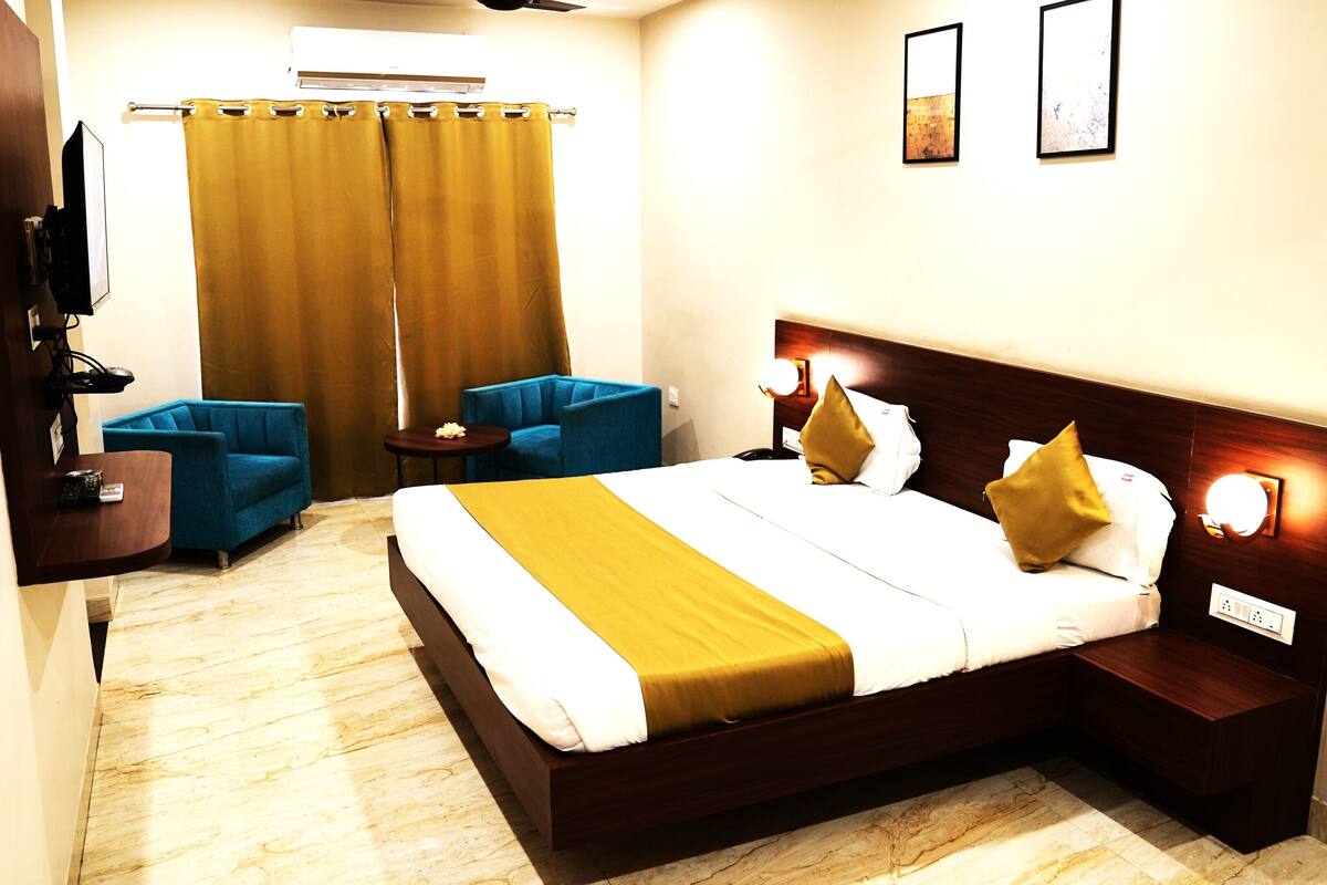 A hotel room with a large bed, two blue armchairs, a small table, a curtain, and two wall-mounted lights.