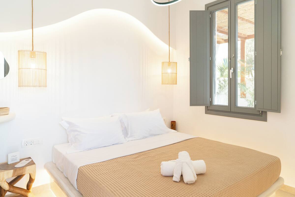 Ein ordentlich bezogenes Bett mit weißer Decke und Kissen, ein Nachttisch aus Holz mit einem Handtuch sowie ein Fenster mit Holzjalousien.