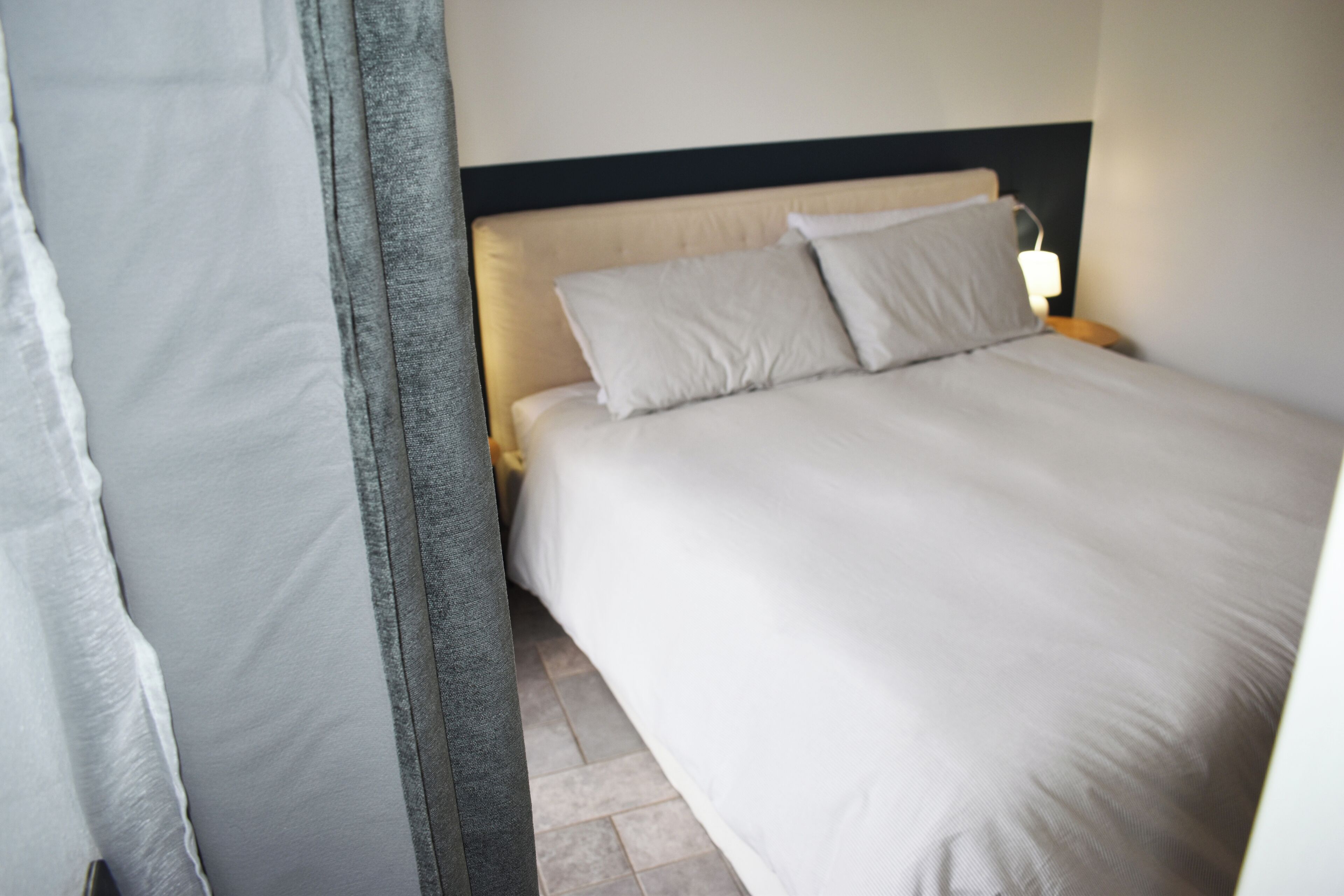 Una cama bien hecha con cabecera, una mesita de noche con lámpara, y una cortina entreabierta.