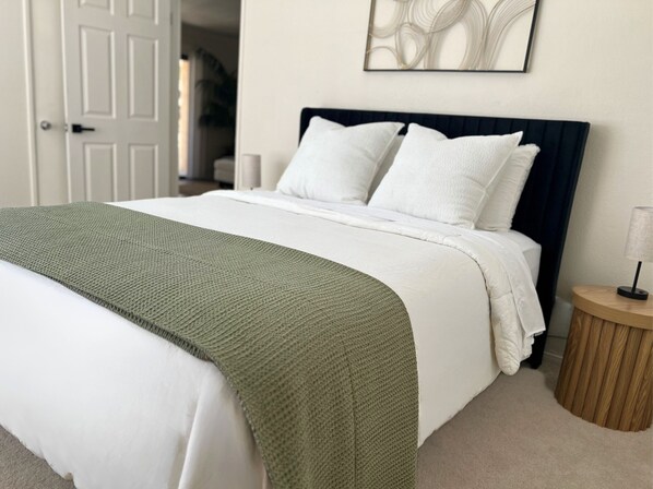 A neatly made bed with white bedding and a green throw blanket, a wooden nightstand with a lamp, and a framed abstract artwork on the wall.